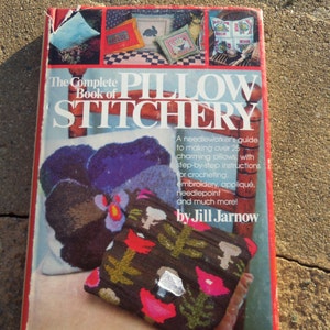May include: The Complete Book of Pillow Stitchery, a book cover featuring a needlework guide to making over 25 charming pillows with step-by-step instructions for crocheting, embroidery, appliqué, needlepoint, and more. The book cover features a colourful pillow with floral designs.