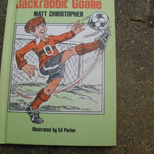 May include: A green book cover with the title "Jackrabbit Goalie" and the author "Matt Christopher". The cover features an illustration of a cartoon boy wearing a red and black soccer uniform, kicking a soccer ball with his right leg. The book is illustrated by Ed Parker.