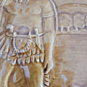 May include: A brown ceramic tile with a raised image of a person wearing a feathered headdress and a skirt. The person is holding a spear in their right hand. The background of the tile is a light brown color.