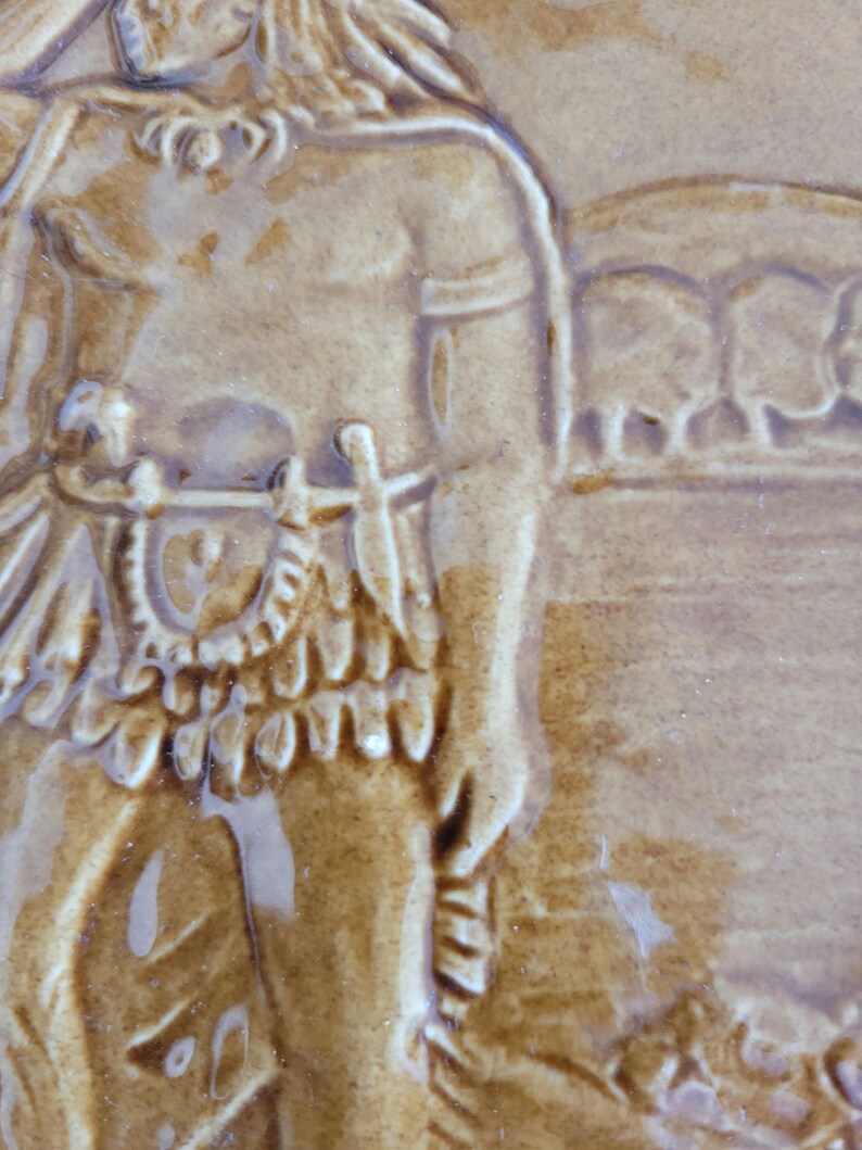 May include: A brown ceramic tile with a raised image of a person wearing a feathered headdress and a skirt. The person is holding a spear in their right hand. The background of the tile is a light brown color.