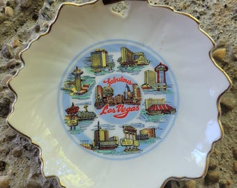 May include: A white porcelain shell-shaped dish with a gold-trimmed scalloped edge. The center features a colorful illustration of various Las Vegas landmarks and casinos, with the red text "Fabulous Las Vegas" printed in the middle of the design.