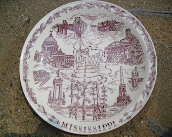 May include: A white ceramic plate with a maroon illustration of Mississippi landmarks, including the New Capitol, Old Capitol, and Mississippi River scenes. The plate has a decorative floral border and the word "Mississippi" at the bottom.