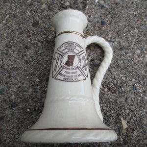 Puede incluir: Taza de cerámica color crema con base acampanada y asa en forma de cuerda. La taza presenta un borde dorado y un gráfico marrón con el texto "63rd Columbia County Volunteer Fireman's Convention 1988". El texto también incluye "Rogers Hose Co. No. 3 Hudson, N.Y."