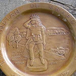 May include: A brown ceramic plate with a raised image of an Indigenous person in a feathered headdress, standing on a bluff overlooking a river. The plate is inscribed with the text "The Indian of Crapo Park, On the Bluffs Above the Mississippi River, Burlington Iowa."