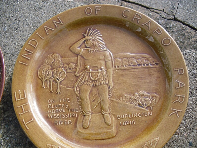 May include: A brown ceramic plate with a raised image of an Indigenous person in a feathered headdress, standing on a bluff overlooking a river. The plate is inscribed with the text "The Indian of Crapo Park, On the Bluffs Above the Mississippi River, Burlington Iowa."
