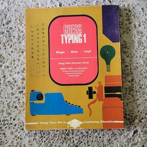 May include: A vintage textbook titled "Gregg Typing 1" with a gold cover and a red rectangular title box. The cover features a blue typewriter illustration and the text "Gregg Adult Education Series".