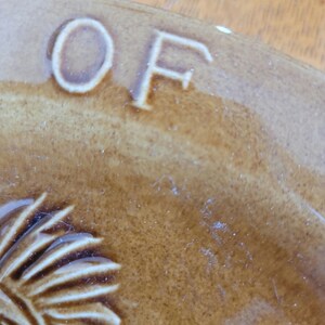 May include: Close-up of a brown ceramic plate with the letters "OF" embossed in white. The plate features a brown and white design of a Native American headdress.