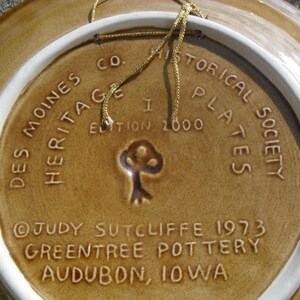 May include: A brown ceramic plate with a white rim and a gold cord attached. The plate has a design of a tree and text that reads "Des Moines Historical Society Plates, I Edition 2000, Heritage, Judy Sutcliffe 1973, Greentree Pottery, Audubon, Iowa."