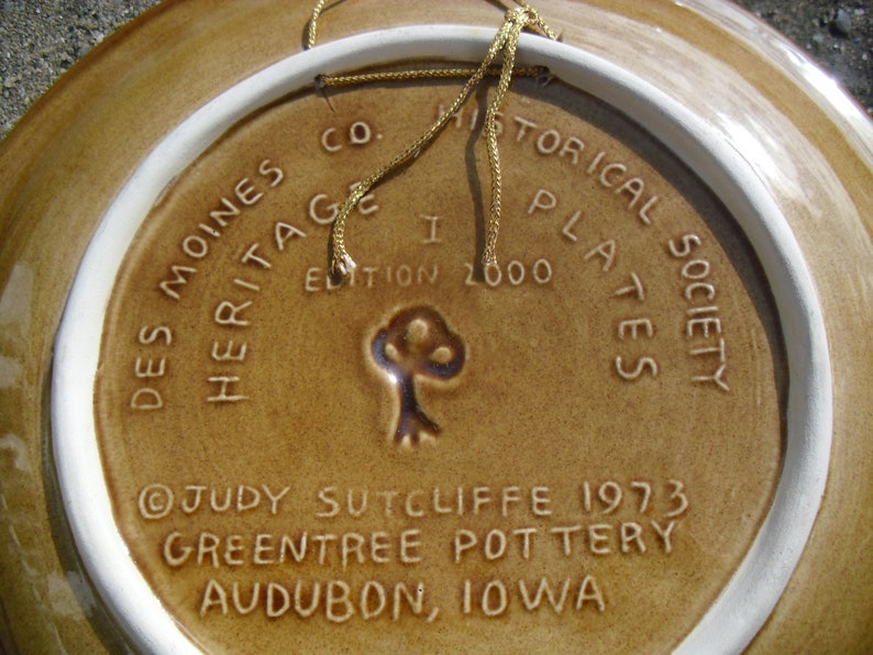 May include: A brown ceramic plate with a white rim and a gold cord attached. The plate has a design of a tree and text that reads "Des Moines Historical Society Plates, I Edition 2000, Heritage, Judy Sutcliffe 1973, Greentree Pottery, Audubon, Iowa."