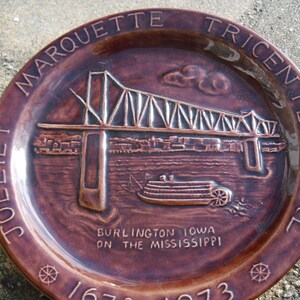 May include: A brown ceramic plate commemorating the Marquette Tricentennial of Burlington, Iowa on the Mississippi. The plate features a raised image of the Burlington  Bridge and a steamboat.
