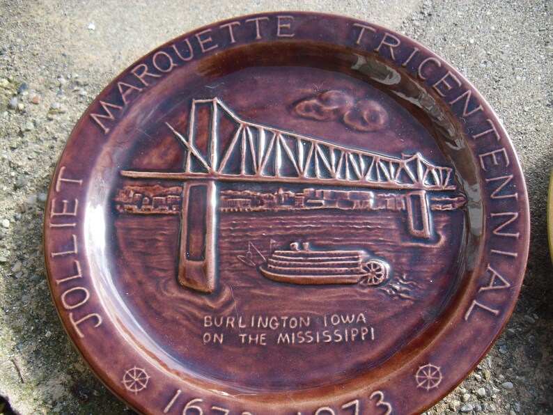 May include: A brown ceramic plate commemorating the Marquette Tricentennial of Burlington, Iowa on the Mississippi. The plate features a raised image of the Burlington  Bridge and a steamboat.