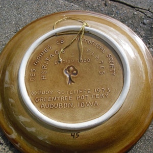 May include: A brown ceramic plate with a white rim and a gold string attached. The back of the plate has white text that reads "Des Moines Co Heritage Historical Plates Society Edition 1000 Judy Sutcliffe 1973 Greentree Pottery Audubon, Iowa" and the number "45".