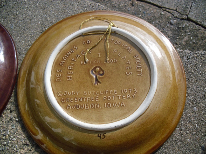 May include: A brown ceramic plate with a white rim and a gold string attached. The back of the plate has white text that reads "Des Moines Co Heritage Historical Plates Society Edition 1000 Judy Sutcliffe 1973 Greentree Pottery Audubon, Iowa" and the number "45".
