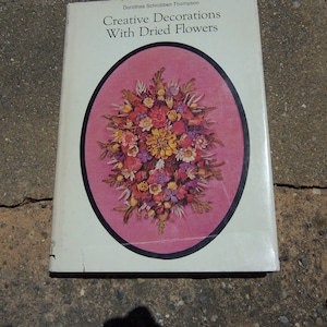 May include: A white book titled "Creative Decorations With Dried Flowers" by Dorothea Schnibbe Thompson. The cover features a pink oval with a floral arrangement of dried flowers.