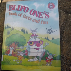 May include: A children's book titled "BLIPO ONE'S book of facts and fun" with colorful illustrations. The cover features a robot character with a red nose and purple glasses, along with cartoon rabbits. The author's signature is visible.