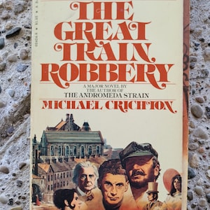 Puede incluir: Un libro de bolsillo vintage titulado The Great Train Robbery de Michael Crichton. La portada de color crema presenta texto rojo en negrita y una ilustración detallada de cinco personas con ropa de época frente a edificios victorianos.