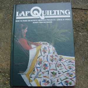 May include: A green book titled "Lap Quilting: How to Make Beautiful Quilted Projects - Large & Small" by Muriel Breckenridge. The cover features a woman sitting in a rocking chair with a colorful quilt.