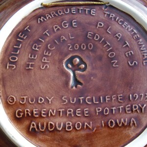 May include: A brown ceramic plate with a white rim. The plate has a gold string attached to the top. The back of the plate has the text "Jolliet Marquette Tricentennial Heritage Plates Special Edition 2000" and "&copy;Judy Sutcliffe 1973 Greentree Pottery Audubon, Iowa".