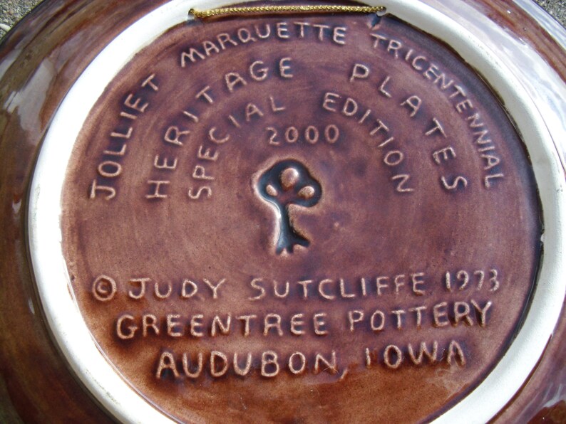 May include: A brown ceramic plate with a white rim. The plate has a gold string attached to the top. The back of the plate has the text "Jolliet Marquette Tricentennial Heritage Plates Special Edition 2000" and "&copy;Judy Sutcliffe 1973 Greentree Pottery Audubon, Iowa".