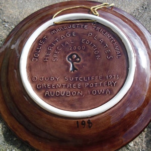 May include: A brown ceramic plate with a white rim. The back of the plate has a brown background with white text that reads "Jolliet Marquette Tricentennial Plates Special Edition 2000" and "Judy Sutcliffe 1973 Greentree Pottery Audubon, Iowa". The number "198" is written in black on the bottom of the plate.