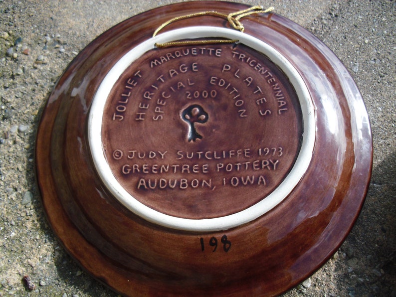 May include: A brown ceramic plate with a white rim. The back of the plate has a brown background with white text that reads "Jolliet Marquette Tricentennial Plates Special Edition 2000" and "Judy Sutcliffe 1973 Greentree Pottery Audubon, Iowa". The number "198" is written in black on the bottom of the plate.