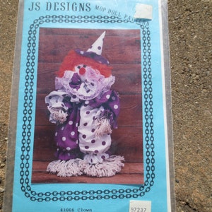 Puede incluir: Un paquete para un patrón de muñeca trapeador de JS Designs. La muñeca es un payaso con peluca roja, lunares blancos sobre tela morada y un cuello de encaje blanco. El paquete es azul con un diseño de cadena.