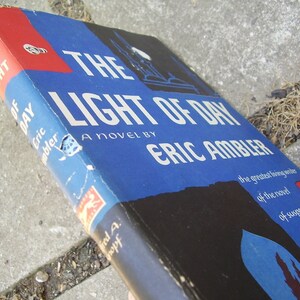 May include: A blue and black paperback book titled "The Light of Day" by Eric Ambler. The book cover features a silhouette of a building and the text "The greatest living writer of the novel of suspense".