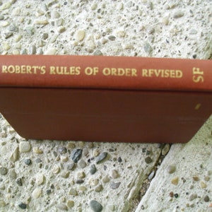 May include: A brown book titled "Robert's Rules of Order Revised" with gold lettering. The book is resting on a textured, gray surface. The book's spine is visible, displaying the title and the initials "SF".