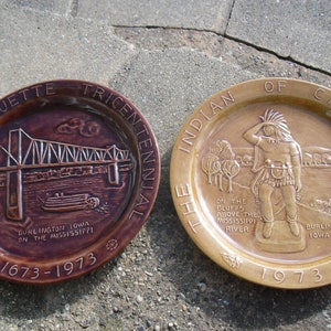 May include: Two ceramic plates commemorating the 1973 tricentennial of the founding of Burlington, Iowa. The first plate features a brown and white image of the Marquette-Joliet Bridge over the Mississippi River. The second plate features a tan image of an indigenous person standing on a bluff overlooking the Mississippi River. Both plates have the text "1673-1973" on them.