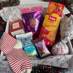 May include: A festive gift box containing a variety of treats. Items include a box of Schar gluten-free Spekulatius biscuits, a bag of Moo Free Snowballs, a red and white striped sock, tea, and chocolate.