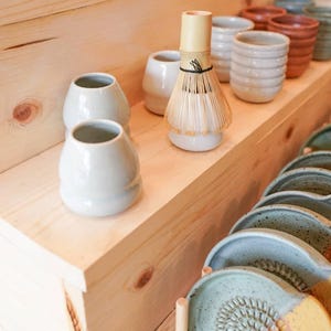 May include: A display of ceramic tea ceremony items. Includes a bamboo whisk, small white ceramic vases, and various textured cups in shades of blue, green, orange, and tan. Also visible are blue and yellow ceramic plates with a grater design.