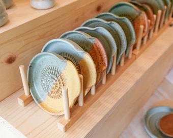 Pode incluir: Exibição de utensílios de cozinha de cerâmica feitos à mão. A exposição inclui uma fileira de pratos raladores em tons de amarelo, azul e laranja, juntamente com pequenas xícaras de cerâmica e um batedor de bambu. Os itens são dispostos em prateleiras de madeira.