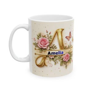 Puede incluir: Taza de cerámica blanca con la letra "A" dorada y el nombre "Amelia" en azul. El diseño incluye rosas rosas, hojas verdes y una mariposa. La taza tiene un asa blanca y un fondo color crema.
