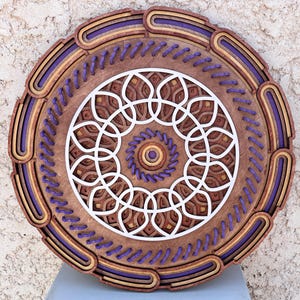 Puede incluir: Una pieza de arte de pared circular de madera con un diseño geométrico en tonos marrones y blancos. El diseño presenta un círculo central con un patrón en espiral, rodeado de una serie de círculos concéntricos y patrones intrincados. El borde exterior de la pieza está decorado con un borde morado y marrón.