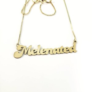 Può includere: Una collana in metallo color oro con un ciondolo a forma di nome in stile calligrafico che riporta la scritta "Melenated" in un carattere corsivo. Il ciondolo è fissato a una sottile catenina a maglie quadrate che sale verso l'alto, con una finitura lucida.