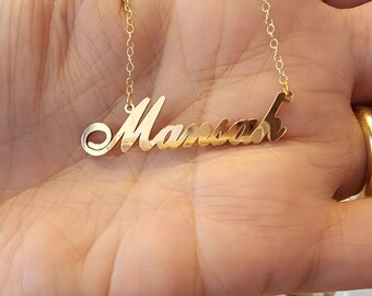May include: A gold-colored name necklace featuring the name Mansah in a cursive script font. The pendant is attached to a delicate gold-colored chain with small circular links, creating a polished and reflective metallic appearance.