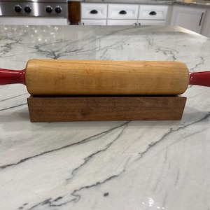 Può includere: Un mattarello di legno con manici rossi su un supporto di legno. Il mattarello è su un piano di lavoro in marmo bianco.