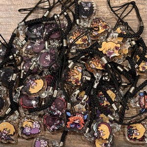 Puede incluir: Una colección de dijes de acrílico transparente con varios personajes ilustrados estilo anime con cabello y atuendos coloridos. Cada dije está unido a un cordón trenzado negro con un conector de metal plateado en un montón.