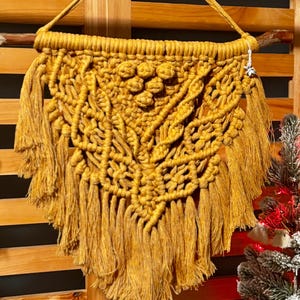 Pode incluir: Uma tapeçaria de macramé amarela mostarda com um padrão geométrico e franjas. A tapeçaria é feita com fibras naturais e tem um estilo rústico e boêmio.
