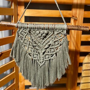 May include: A green macrame wall hanging with a geometric pattern and fringe. The hanging is attached to a wooden dowel.