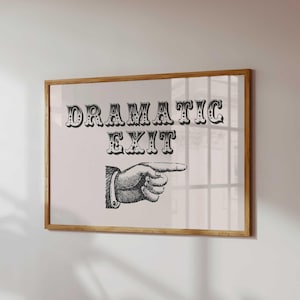 May include: A framed print featuring the words "DRAMATIC EXIT" in decorative black font. Beneath the text is a black and white illustration of a hand pointing right. The frame is made of light-coloured wood.