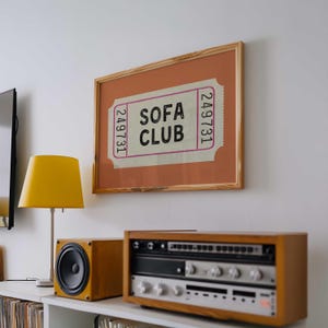 May include: A framed print with the text "SOFA CLUB" on a ticket design. The print is in a wooden frame and hangs above a stereo system and a yellow lamp. The stereo system has a wooden cabinet.