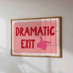 May include: A framed art print featuring the words "DRAMATIC EXIT" in red, with a pink hand pointing right. The background is a light pink rectangle with a textured, brushstroke effect. The frame is wooden.
