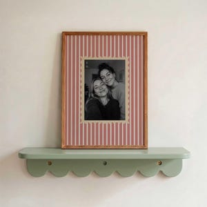 May include: A rectangular wooden picture frame containing a black and white photograph of two people. The frame features a vertical red and white striped mat with a scalloped inner border. The frame rests on a sage green scalloped wall shelf.