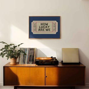 May include: A framed print with the text "HOW LUCKY ARE WE" in a ticket design. The print is displayed above a wooden cabinet with a record player, vinyl records, and a potted plant. The cabinet has a mid-century modern design.