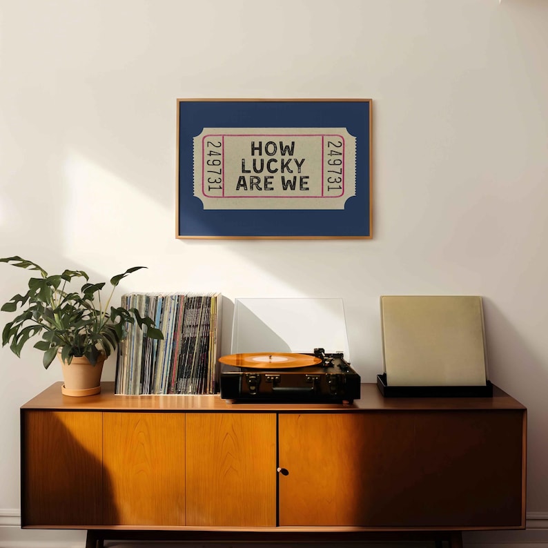 May include: A framed print with the text "HOW LUCKY ARE WE" in a ticket design. The print is displayed above a wooden cabinet with a record player, vinyl records, and a potted plant. The cabinet has a mid-century modern design.
