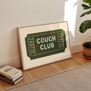 Peut inclure: Une affiche encadrée avec un design de billet vert affichant "COUCH CLUB" en blanc. Le billet porte le numéro "249731" de chaque côté. L'affiche est appuyée contre un mur blanc, avec des livres et une plante en pot à proximité.