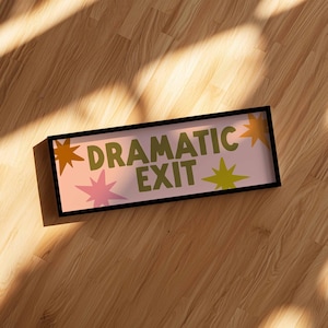 May include: A rectangular sign with a black frame featuring a light pink background and the text "DRAMATIC EXIT" in olive green letters. The design includes several multi-pointed star shapes in shades of orange, pink, and lime green.