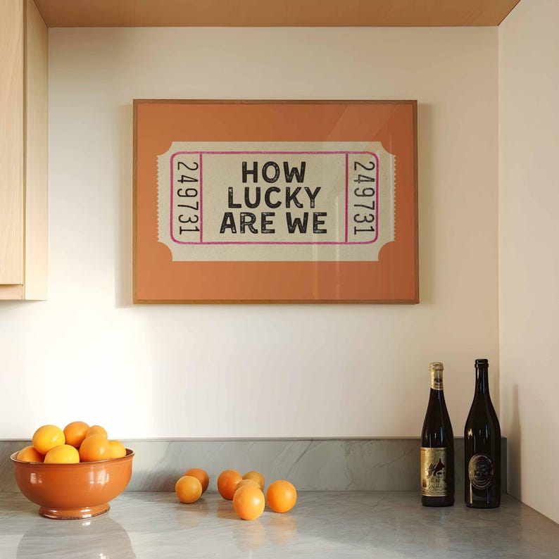 May include: A framed print with the text "HOW LUCKY ARE WE" in a ticket design. The print is displayed above a countertop with a bowl of oranges and two wine bottles. The print has a peach-colored background and a wooden frame.