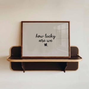 'How Lucky Are We' print. Available in 8 different colour options. The poster in the picture has a beige background with the words 'how lucky are we' in small cursive beige font in the centre, with a small 4 leaf clover shamrock just beneath it. Displayed in a horizontal wooden frame sitting on a black retro wooden shelf.